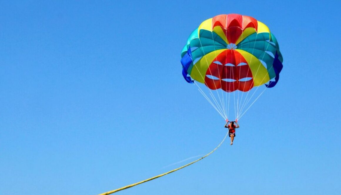 What is parasailing: person soaring under a vibrant colorful parasail against a clear blue sky.