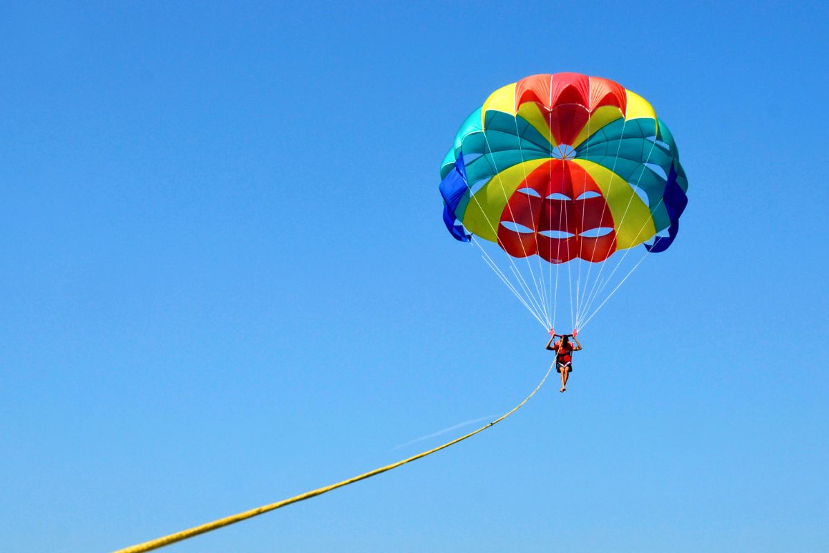 What is parasailing: person soaring under a vibrant colorful parasail against a clear blue sky.
