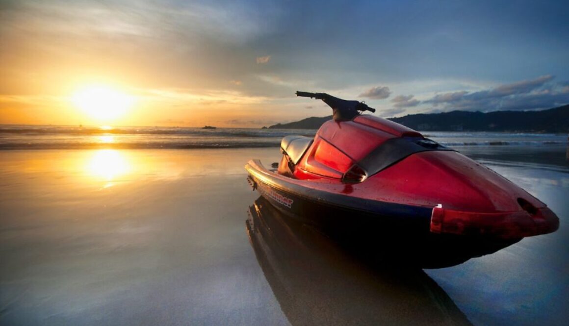 Red personal watercraft parked on a sandy beach at golden sunset with dramatic clouds and calm ocean in the background, capturing the serene end of a day on the water.