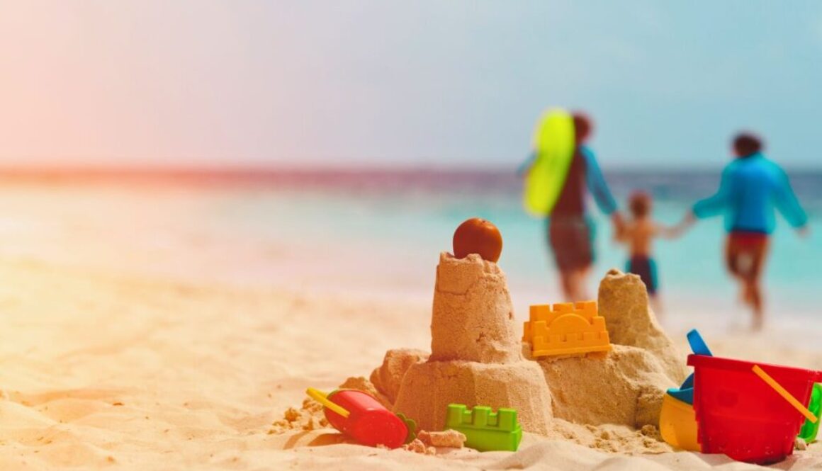 Colorful sandcastle built on a sunny beach with toy shovel, bucket, castle mold, and popsicle shapes, topped with an orange ball, with a happy family walking hand-in-hand in soft focus along the shoreline, capturing joyful moments of family travel