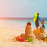 Colorful sandcastle built on a sunny beach with toy shovel, bucket, castle mold, and popsicle shapes, topped with an orange ball, with a happy family walking hand-in-hand in soft focus along the shoreline, capturing joyful moments of family travel