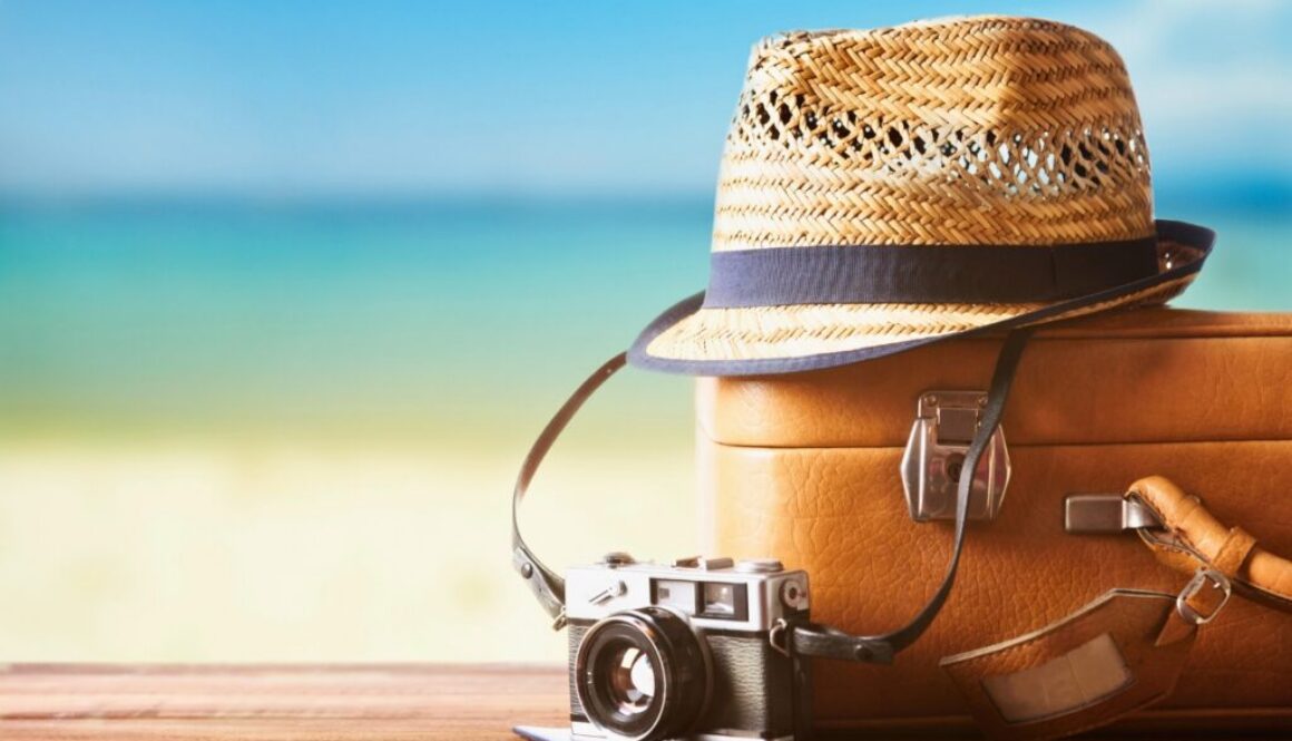 Vintage suitcase, straw hat, and retro camera on a wooden deck by the beach, representing essentials for deciding where to travel in summer
