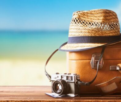 Vintage suitcase, straw hat, and retro camera on a wooden deck by the beach, representing essentials for deciding where to travel in summer