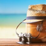 Vintage suitcase, straw hat, and retro camera on a wooden deck by the beach, representing essentials for deciding where to travel in summer