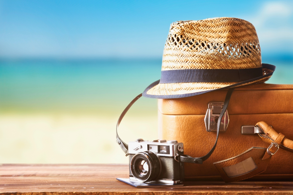 Vintage suitcase, straw hat, and retro camera on a wooden deck by the beach, representing essentials for deciding where to travel in summer