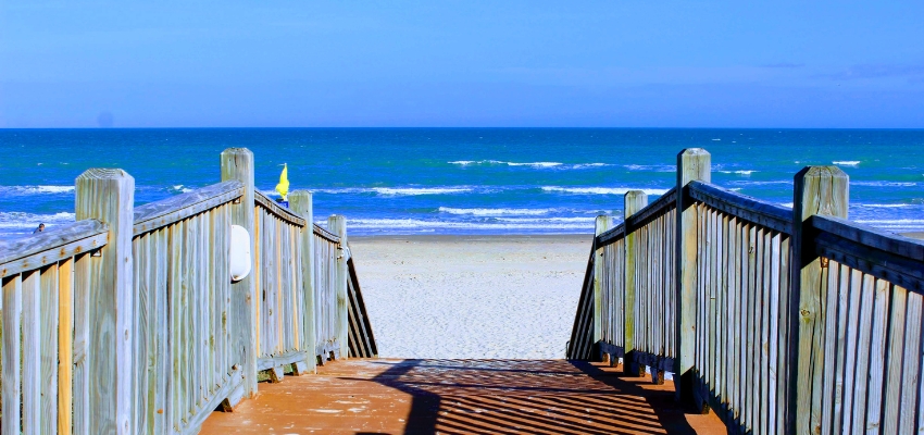 Wooden boardwalk leading down to a sunny ocean beach with blue skies, capturing the ideal summer getaway for travelers deciding where to travel in summer